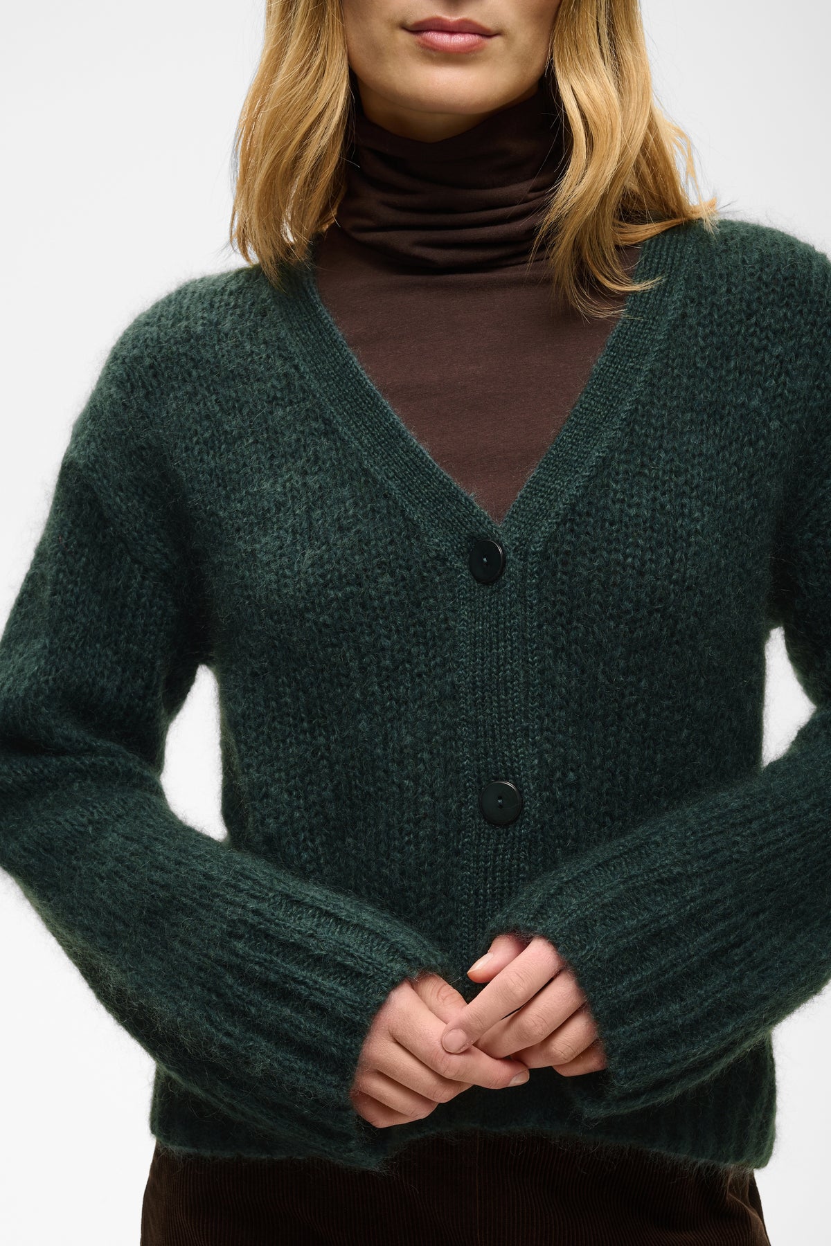 A woman wearing a dark forest green ribbed knit cardigan sweater over a dark brown turtleneck, shown from the chin down against a plain light background.
