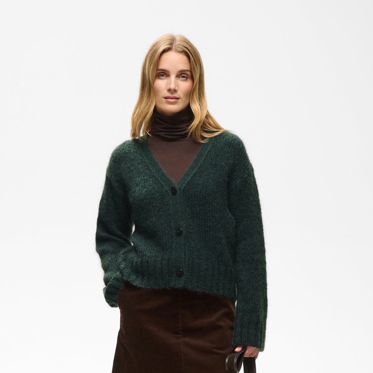 A woman stands against a light gray studio background wearing a dark green fuzzy knit cardigan over a dark brown turtleneck, a dark brown corduroy skirt, black knee-high boots, and holding a black tote bag.