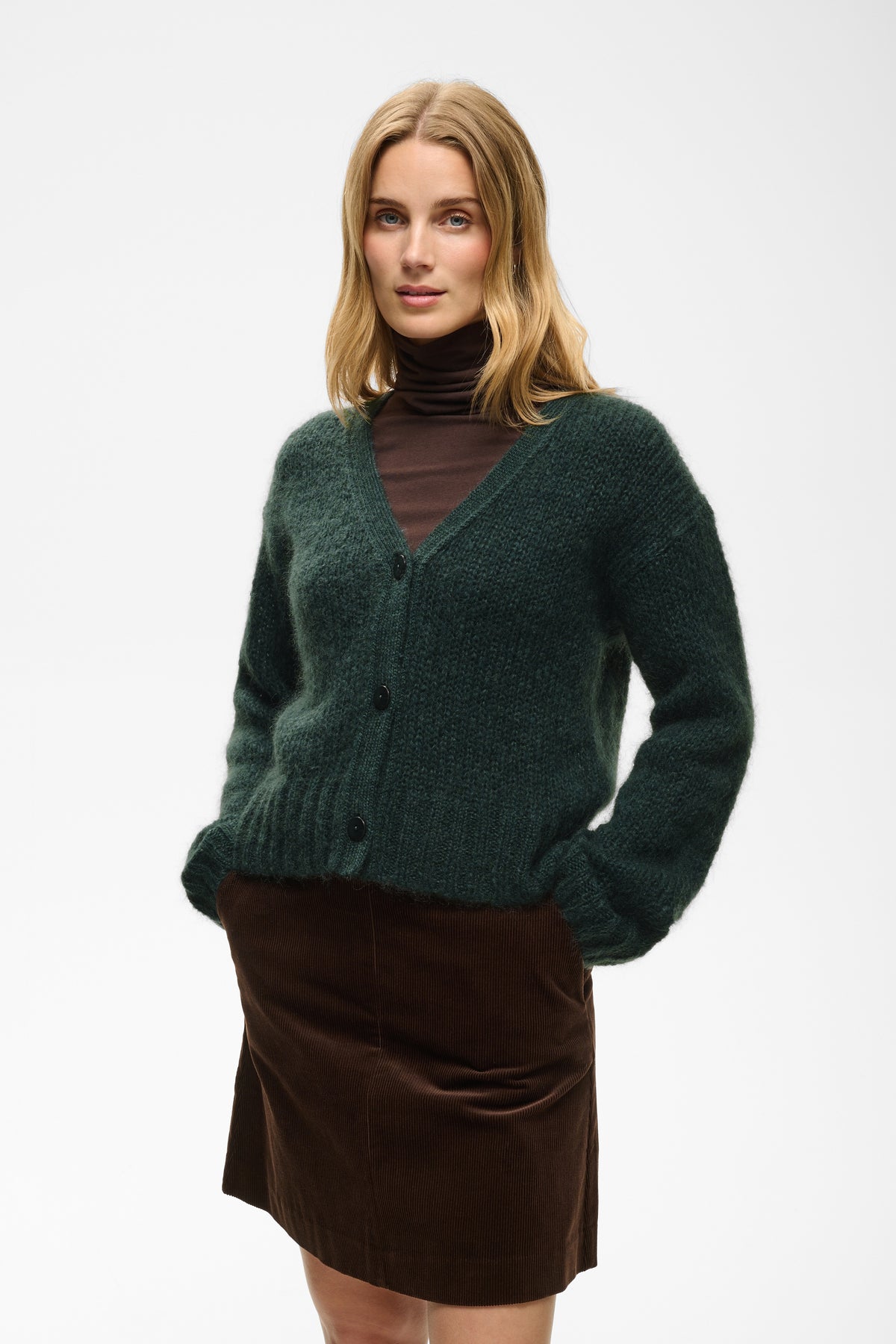 A woman modeling a dark green fuzzy button-up cardigan sweater over a dark brown turtleneck, paired with a brown skirt, against a plain white studio background.