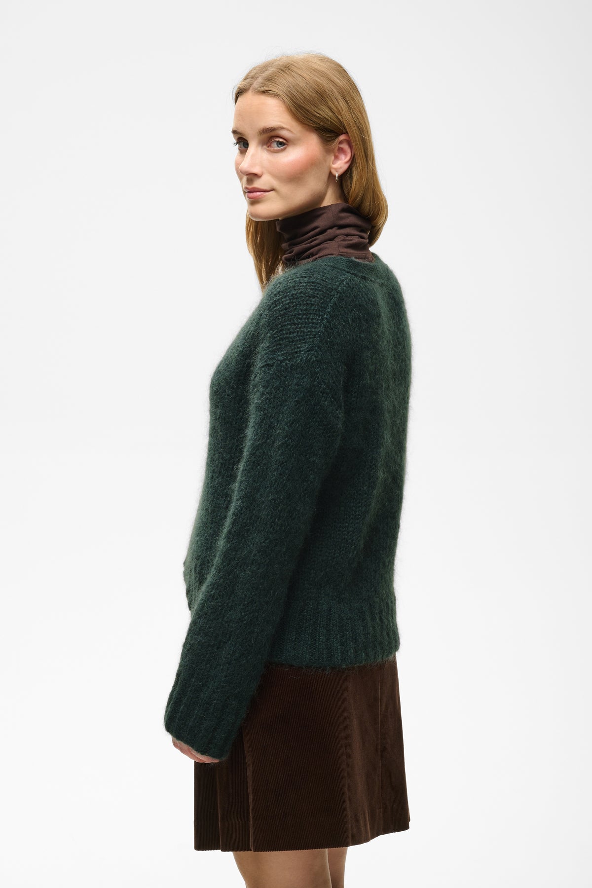 A woman shown in side profile wearing a dark green fuzzy knit sweater over a dark brown mock-neck top and a brown skirt, against a plain white studio background.