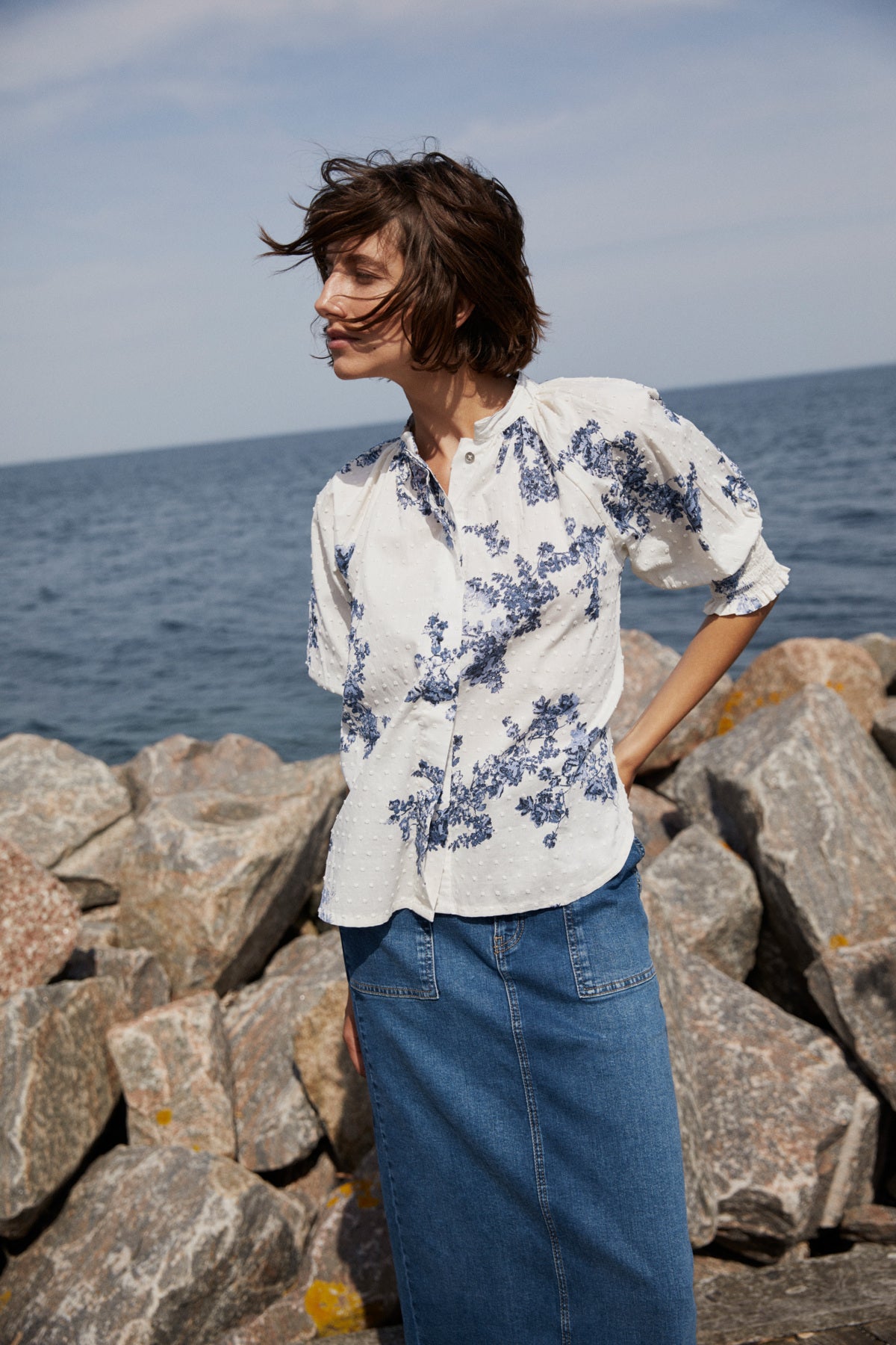 Woman standing on rocks by the sea wearing a floral blouse and jeans.