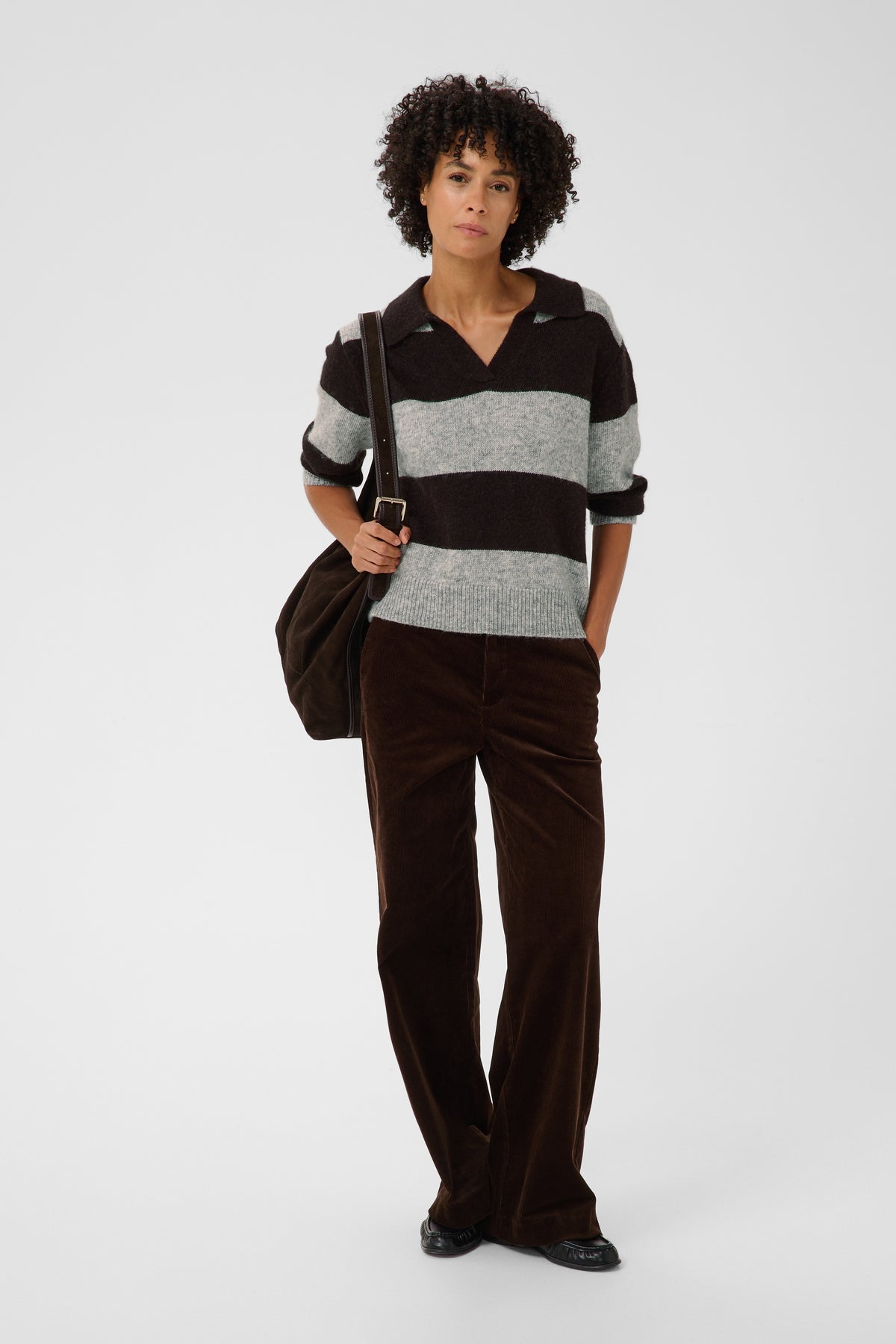 A model stands against a light gray studio background wearing a dark brown and heather gray striped polo sweater, dark brown corduroy trousers, and black shoes, while carrying a dark brown shoulder bag.