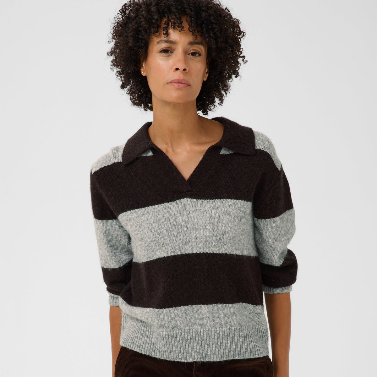 A model with curly black hair wearing a dark brown and light gray striped knit polo sweater and dark brown corduroy trousers, photographed against a light gray studio background.