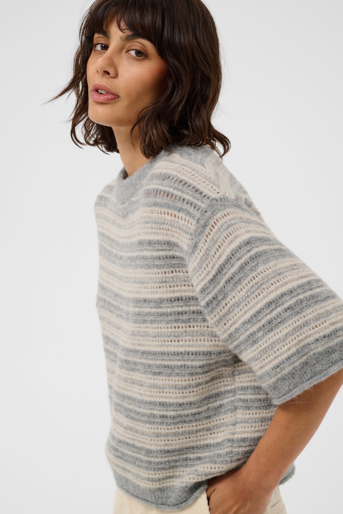 A woman wearing a fuzzy knit short-sleeve sweater with horizontal gray and off-white stripes, photographed against a clean light gray studio background.