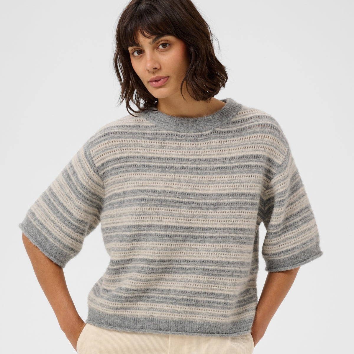 A woman wearing a gray and beige horizontally striped knit sweater with elbow-length sleeves, paired with cream-colored pants, against a plain light gray studio background.
