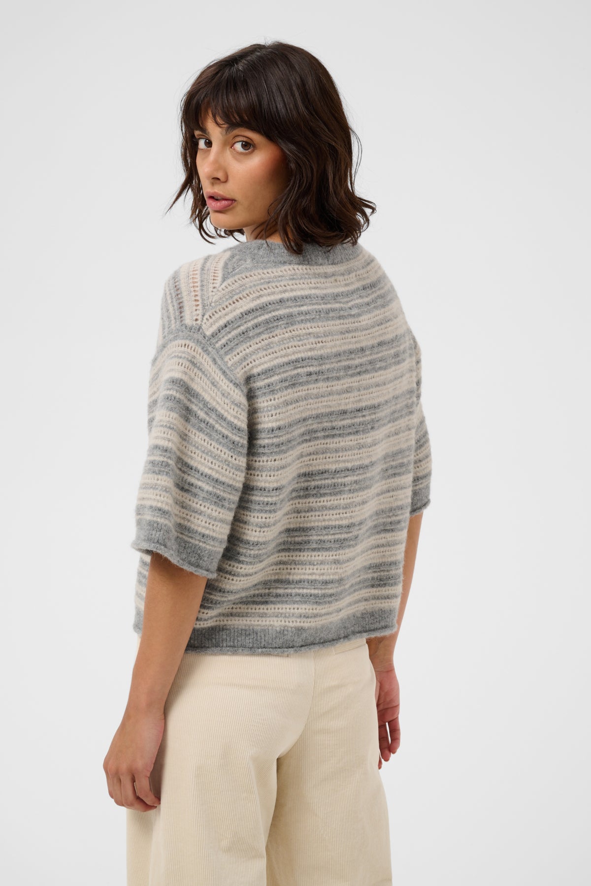 A woman wearing a gray and beige striped knit sweater with elbow-length sleeves, shown from a three-quarter back view against a plain light gray studio background.