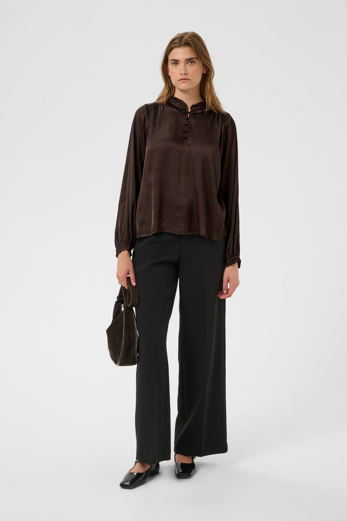 A model stands against a light gray studio background wearing a dark brown long-sleeve blouse with a gathered neckline and black wide-leg trousers, holding a dark brown handbag.
