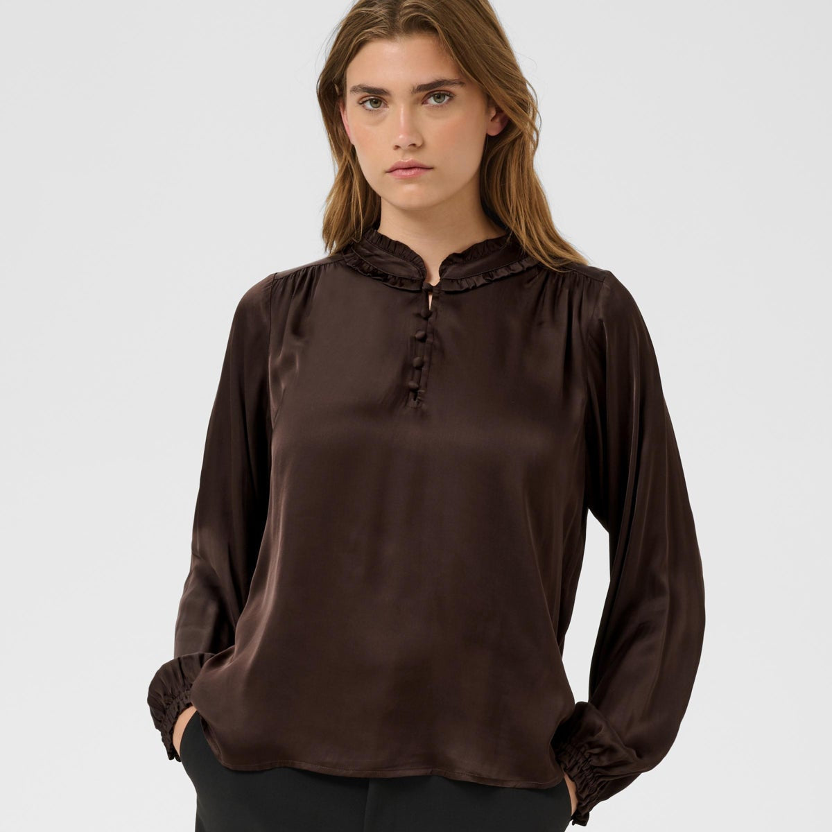 A woman modeling a dark brown long-sleeve blouse with a ruffled mock neck and button placket, paired with black trousers, against a light gray studio background.