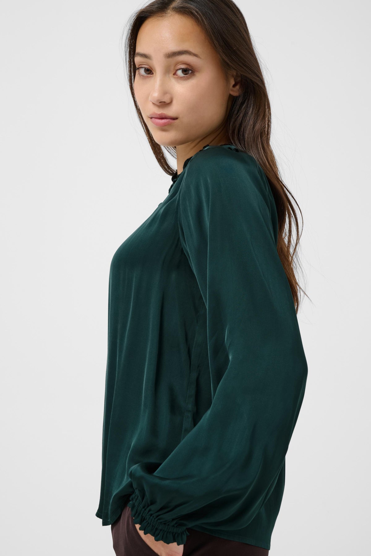 A woman wearing a dark green long-sleeve blouse with a gathered, ruffled hem, shown from a side-back angle against a light gray studio background.