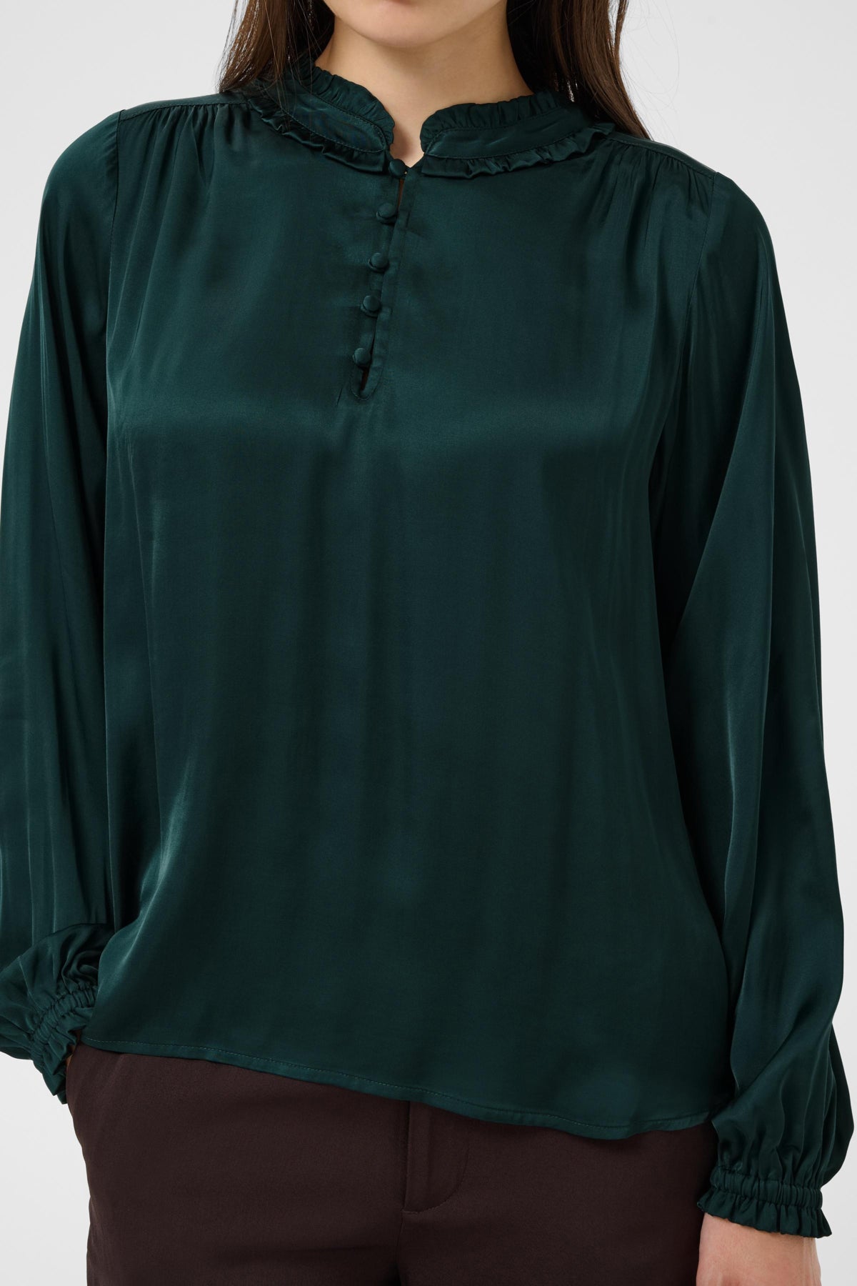 A dark green satin blouse with a ruffled mock neck, buttoned placket, and gathered long sleeves, shown on a model against a light gray background.