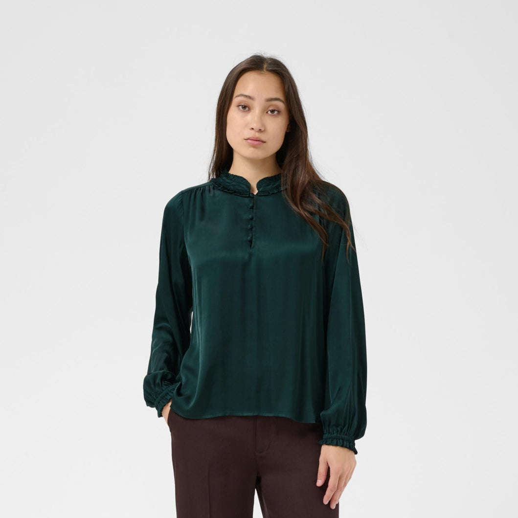 A woman modeling a dark green long-sleeve satin blouse with a high collar and front buttons, paired with dark brown wide-leg trousers, against a light gray studio background.
