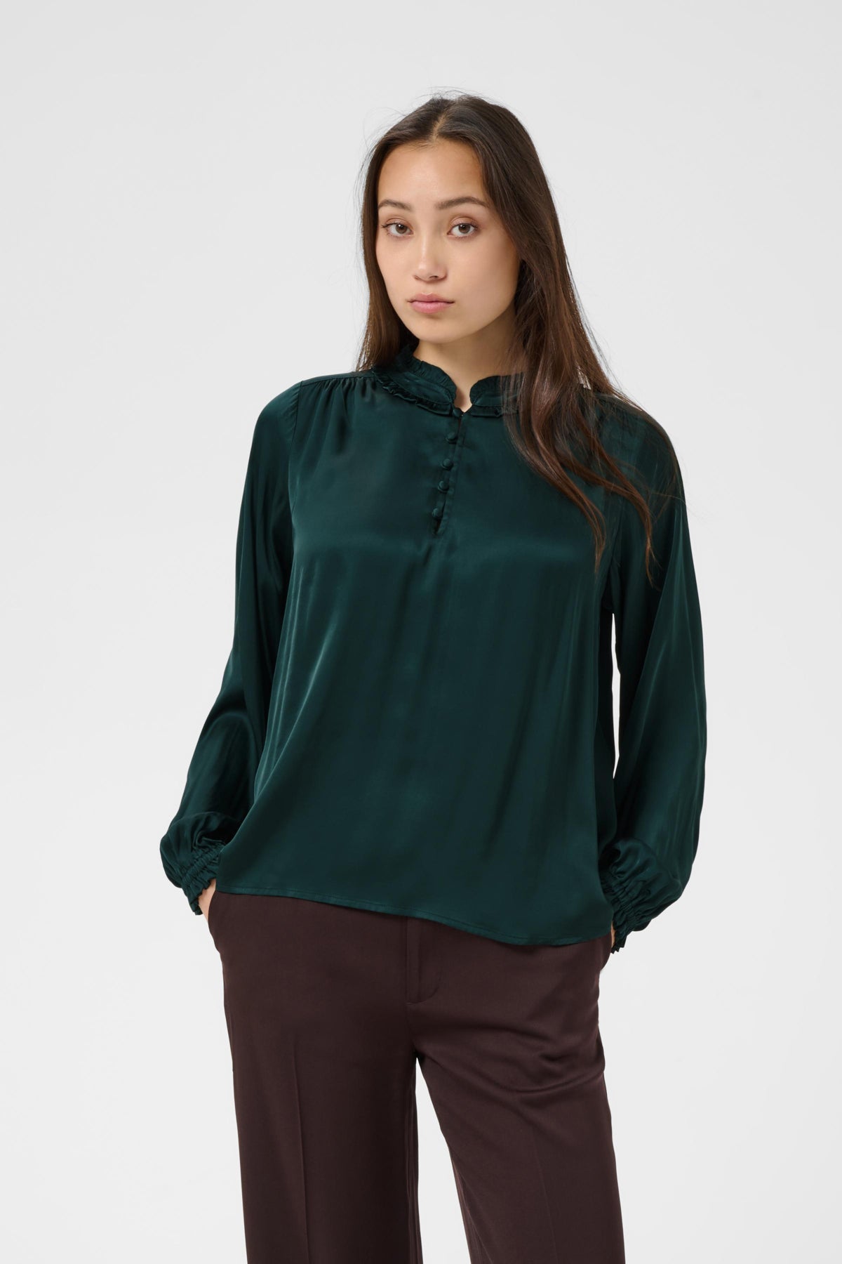 A woman modeling a dark green long-sleeve blouse with a gathered mock neck and button placket, paired with dark brown pants, against a light gray studio background.