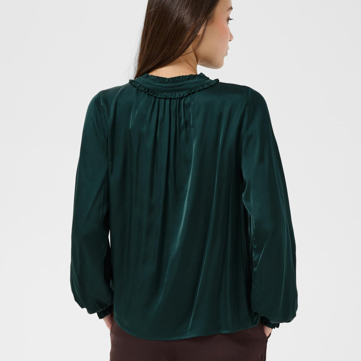 A woman shown from the back wearing a dark green long-sleeve blouse with a gathered, pleated neckline and dark brown trousers, against a light gray studio background.