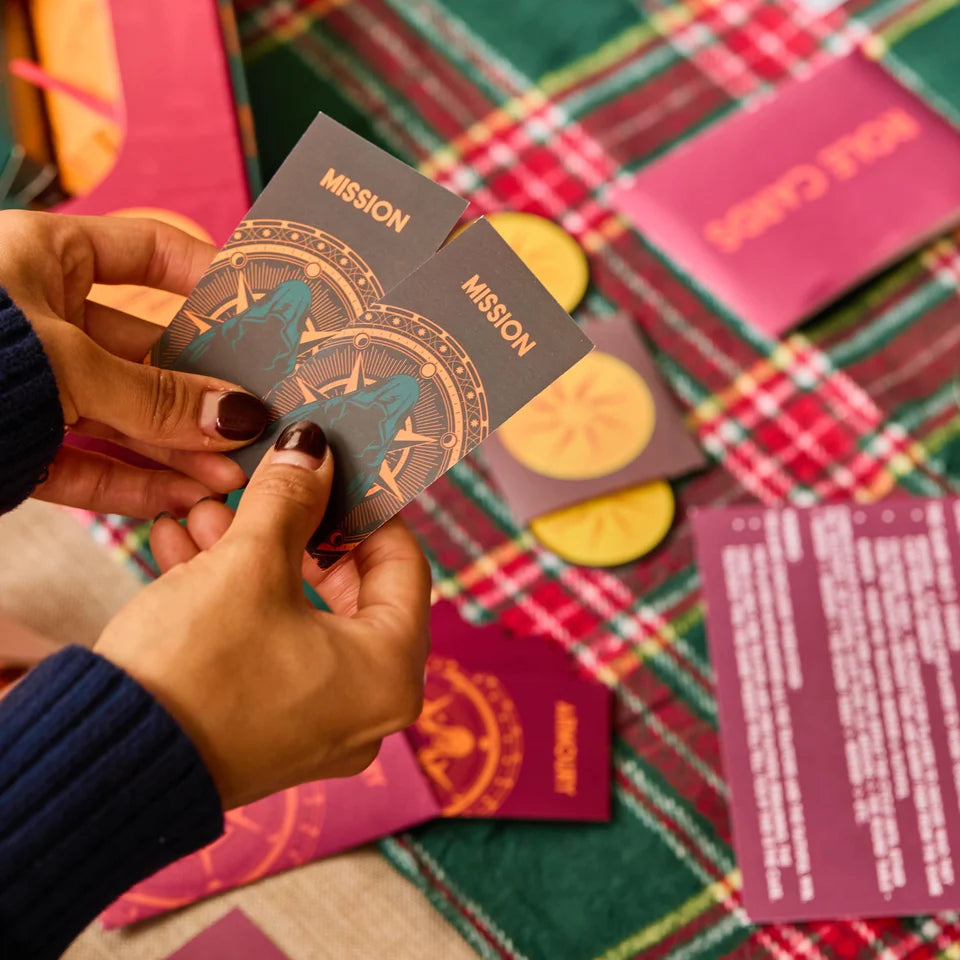 A close-up of hands holding two printed cards labeled "MISSION" over a red, green, and white plaid blanket, with additional matching cards and printed inserts scattered around.