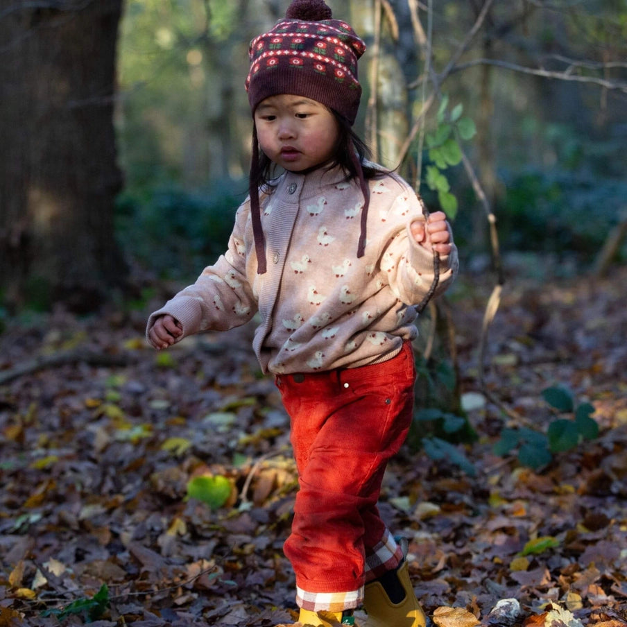 Child in a forest wearing a patterned hat, light-colored jacket, and red pants.