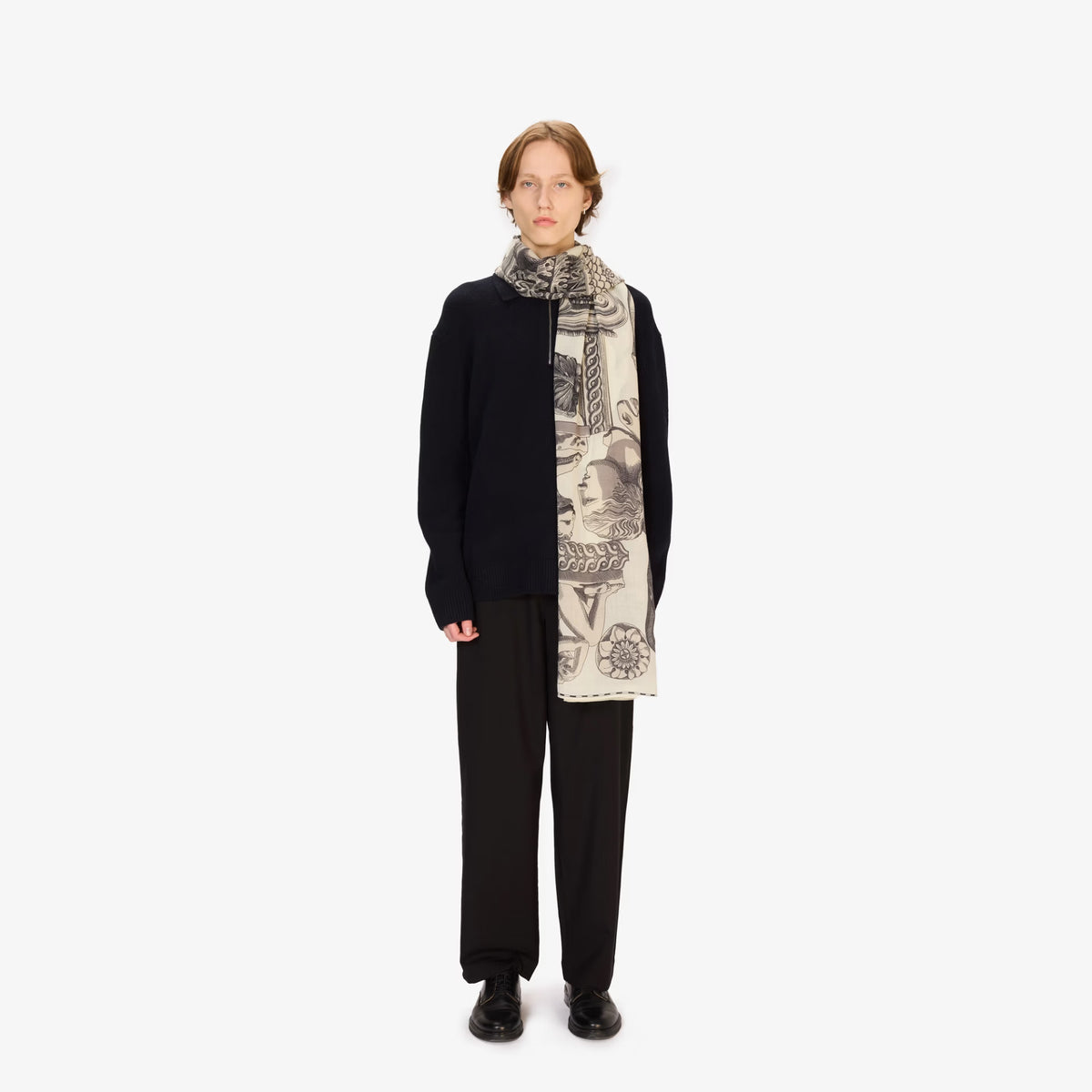 A model stands against a plain white studio background wearing a black cardigan, black trousers, black shoes, and a cream scarf with a detailed black graphic print.