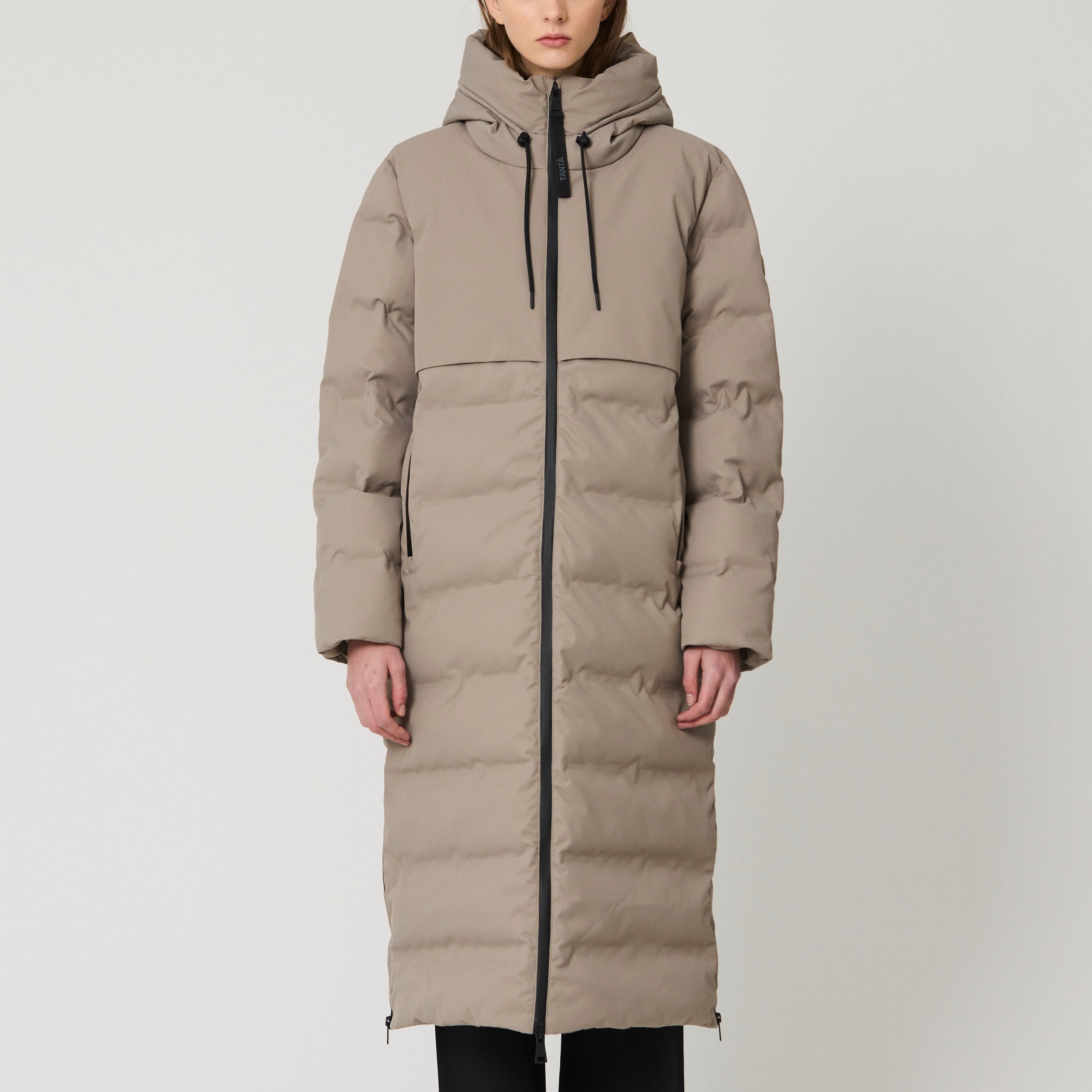 A model stands against a light gray studio background wearing a long, hooded puffer coat in a warm taupe-beige color, paired with black wide-leg pants and brown shoes.