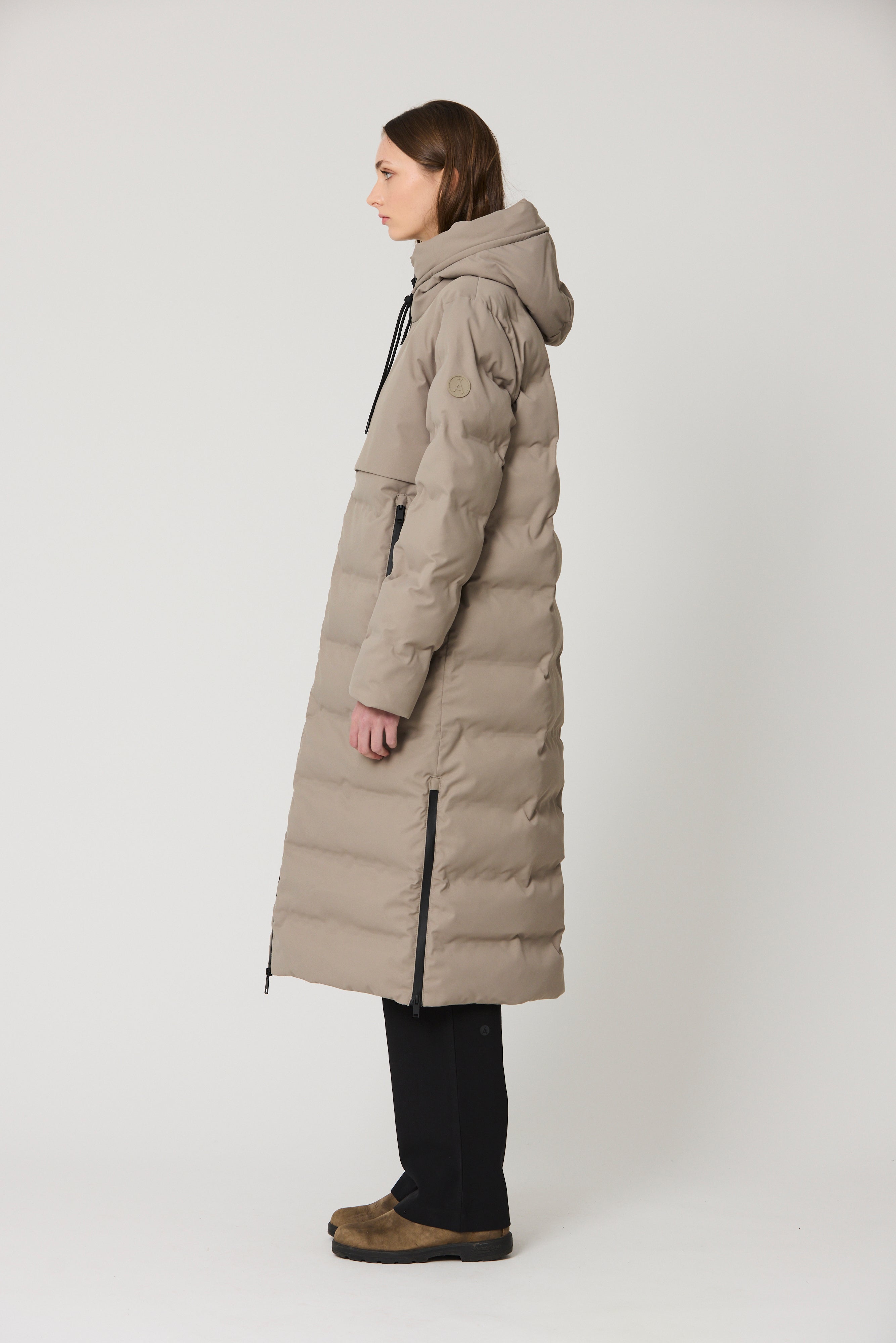 A woman in profile wearing a long, light taupe-beige hooded puffer coat with black zipper details, paired with black pants and brown shoes, photographed against a neutral light gray studio background.