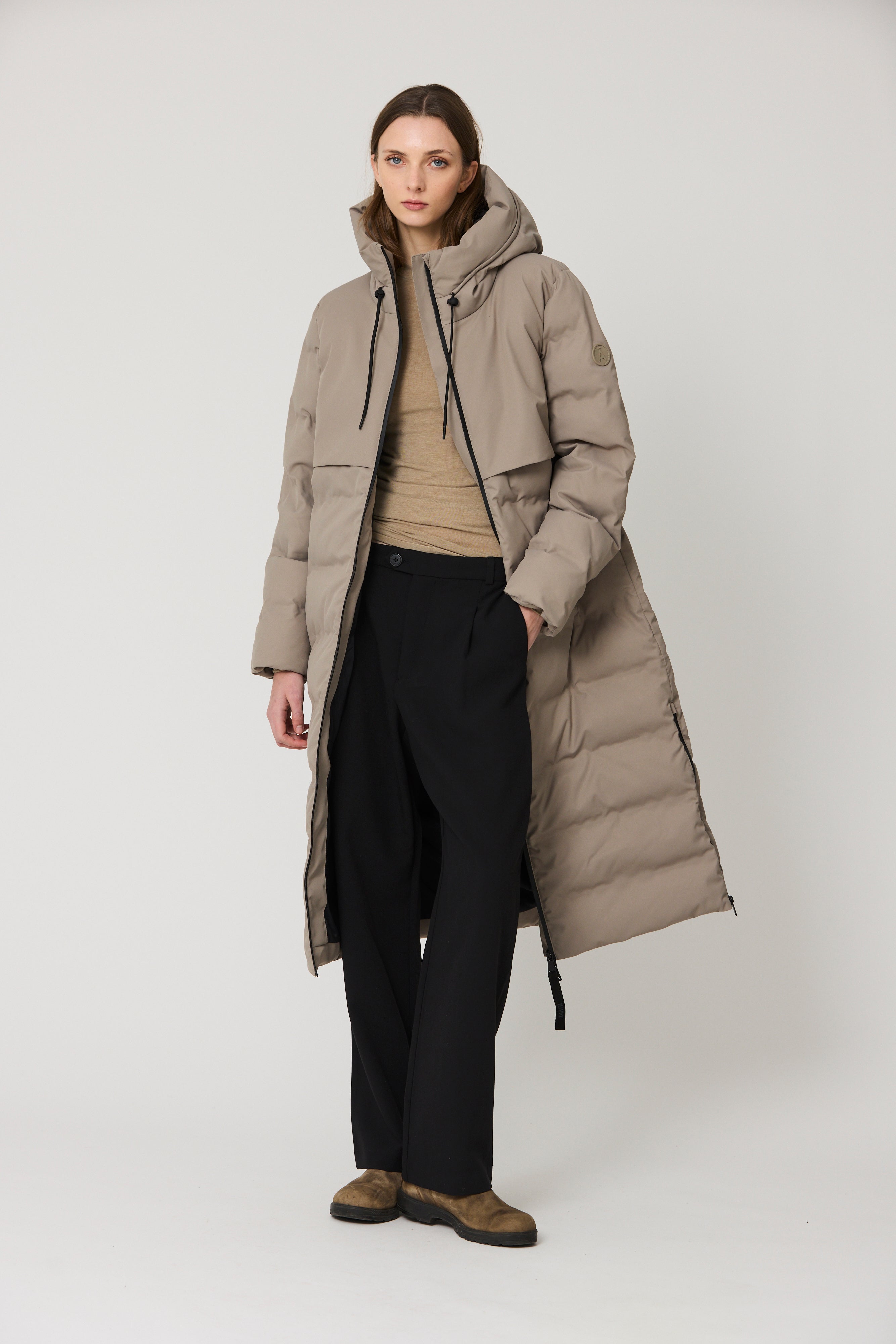 A model stands against a light gray studio background wearing a long taupe quilted puffer coat over a beige top, black trousers, and brown shoes.