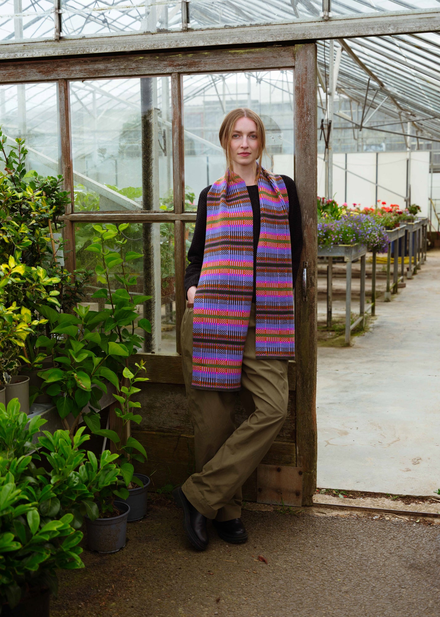 A woman stands in a greenhouse doorway wearing a long, multicolored striped knit scarf over a black top and olive pants.
