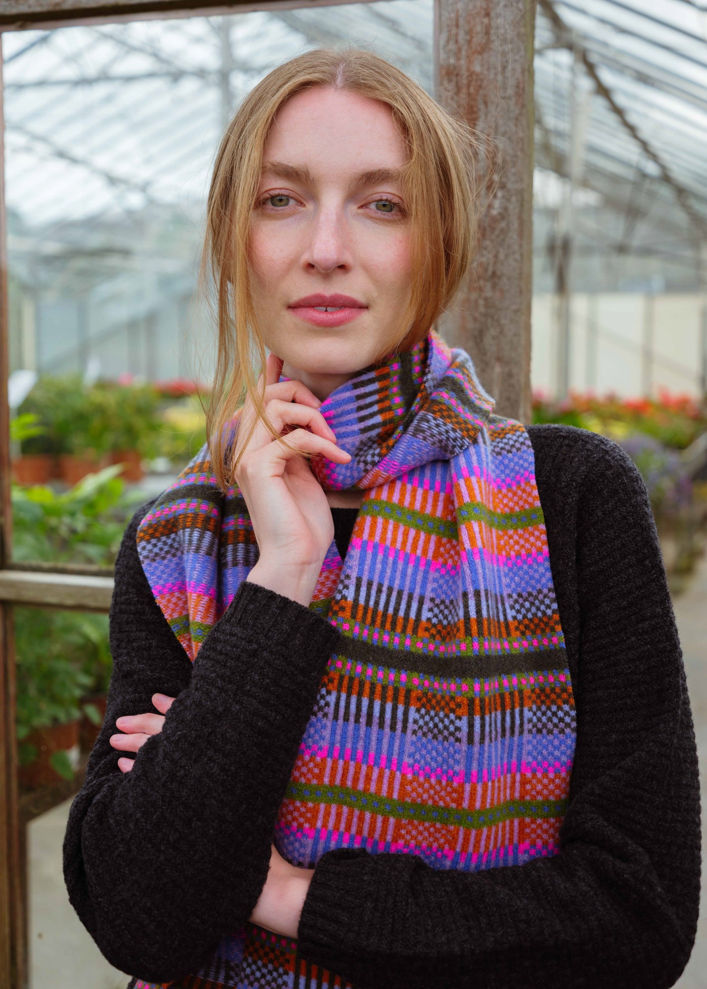 A woman wearing a black knit sweater and a vibrant, multicolored patterned scarf with purple, blue, pink, orange, and green stripes, standing inside a greenhouse.
