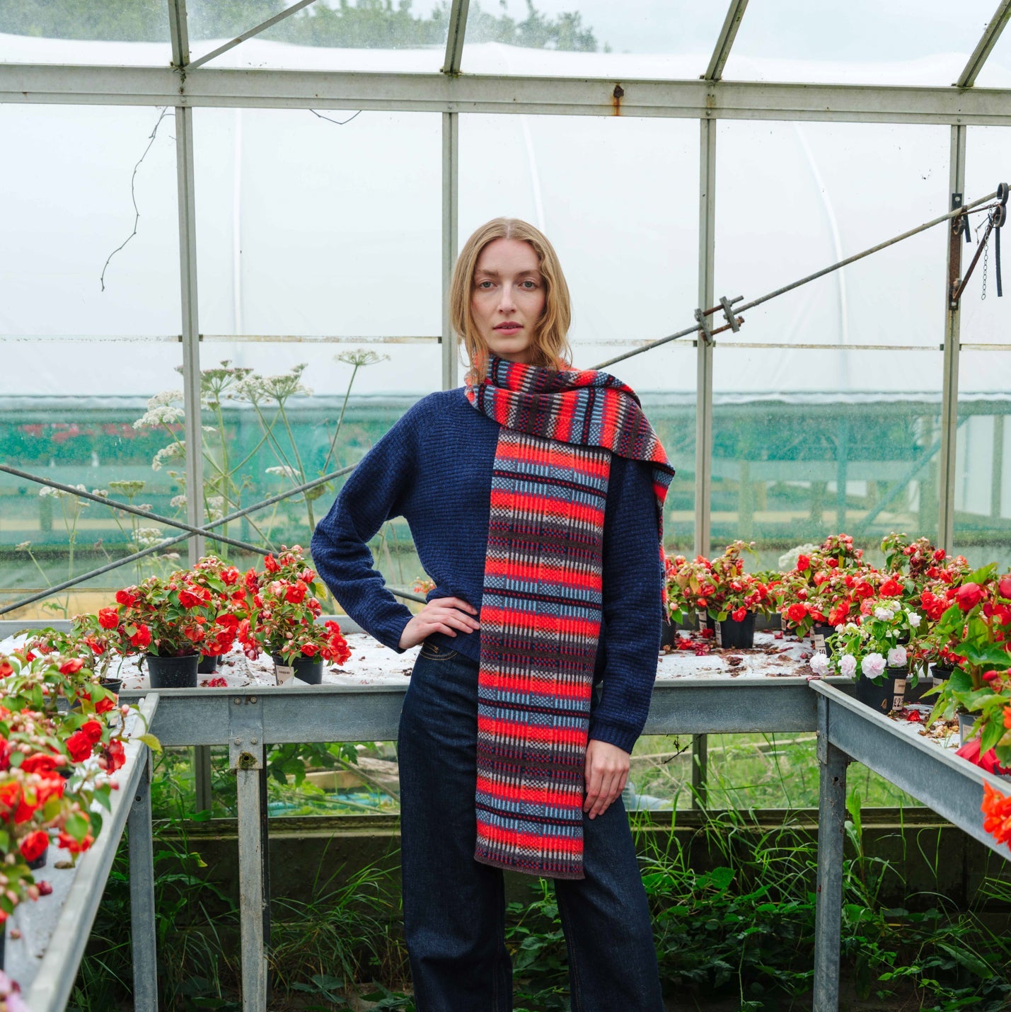 A woman stands inside a greenhouse wearing a navy blue sweater, wide-leg navy pants, a red and navy striped knit scarf, and red shoes. Flower beds and metal tables are visible around her.