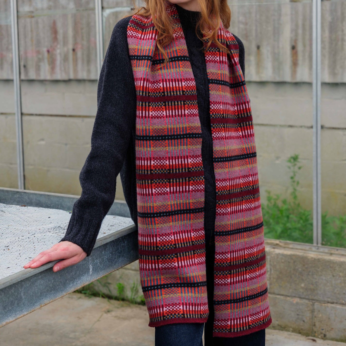 A woman with red hair stands outdoors in front of a greenhouse, wearing a long black cardigan with a red, pink, and orange patterned knit wrap or vest draped over her shoulders.