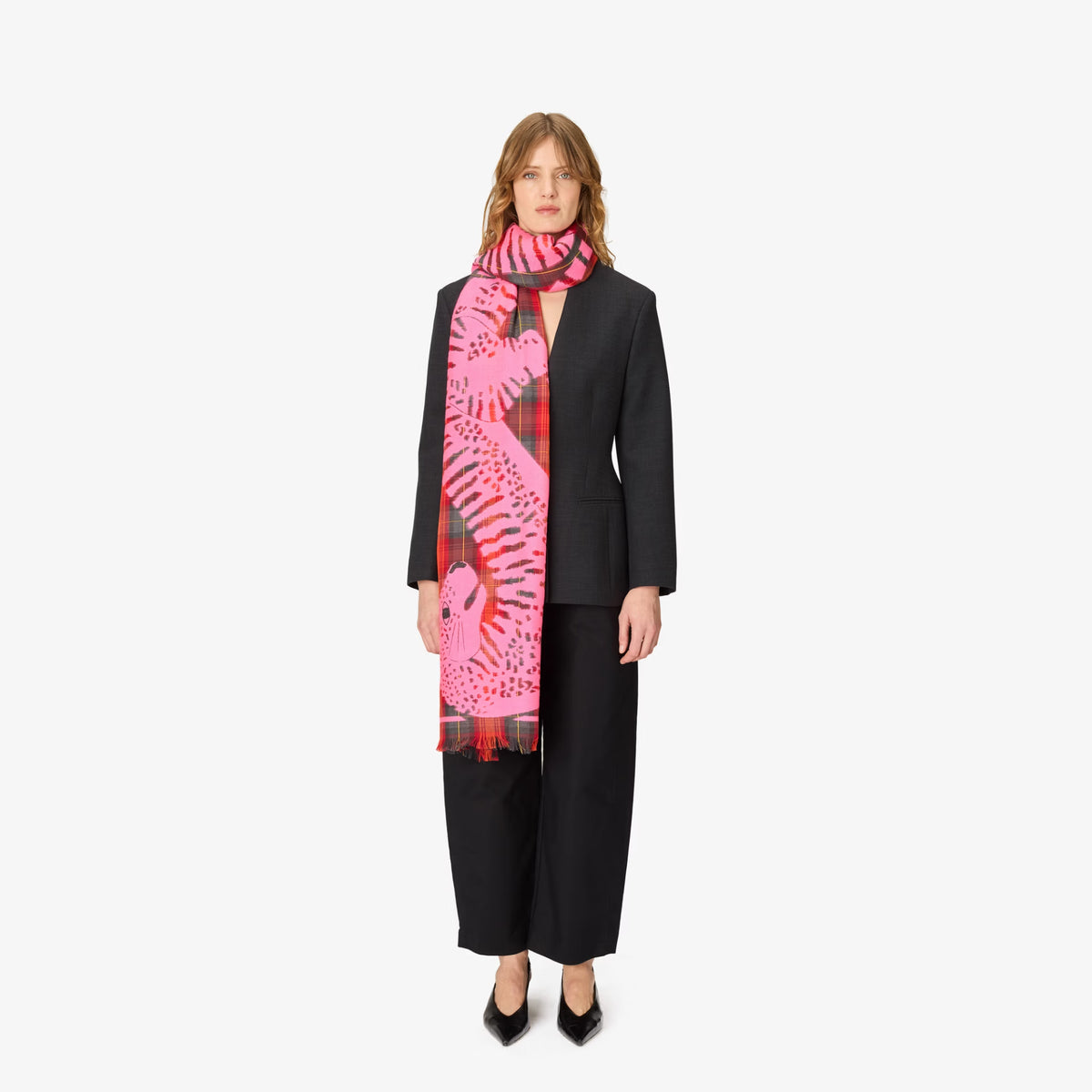 A model wearing a black tailored suit with a vibrant pink scarf featuring a bold leopard print and red plaid accents, posed against a solid off-white background.