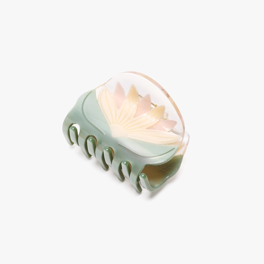 A translucent resin claw hair clip featuring a lotus flower design in pale pink and cream, with a sage green base, shown on a light gray background.