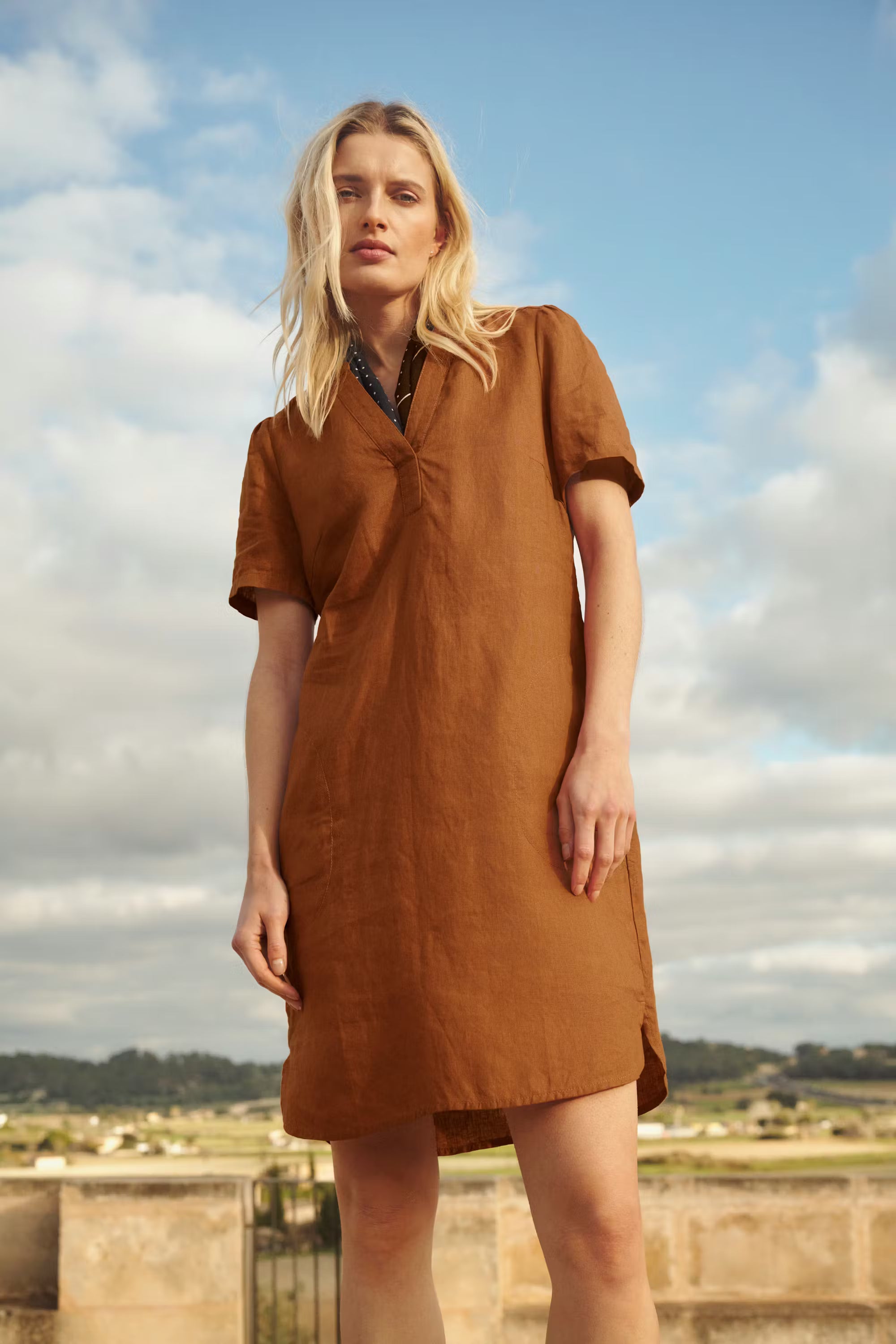 Woman wearing a brown dress standing outdoors with a blue sky and clouds in the background