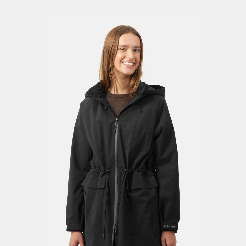 Smiling woman in a black, hooded, zip-up IISE JACOBSEN parka with drawstring waist.