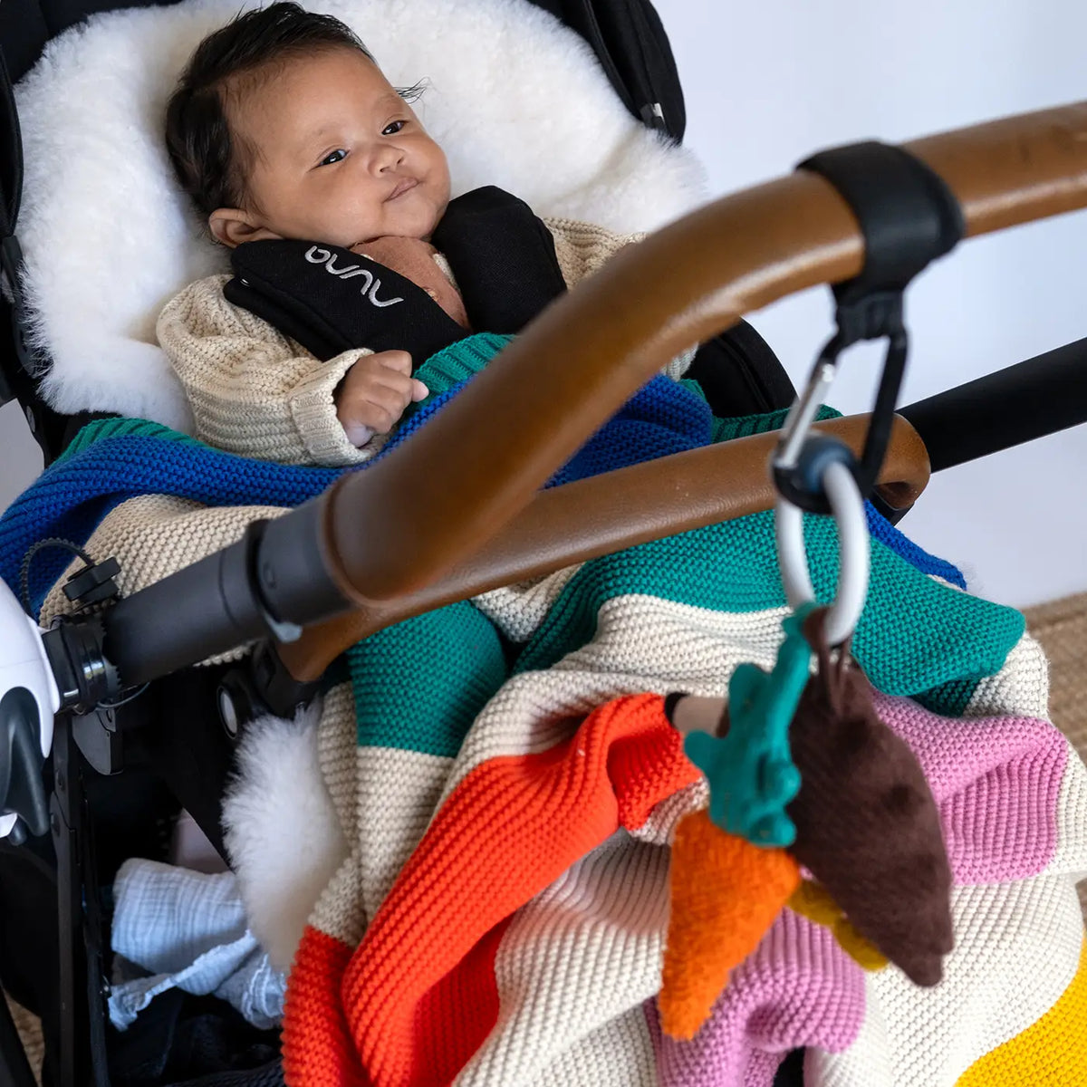 A baby wearing a cream knit sweater and black harness lies in a stroller, wrapped in a chunky knit blanket with bold stripes of teal, orange, pink, and cream. A brown stroller handlebar and a colorful plush carrot toy hang in front.