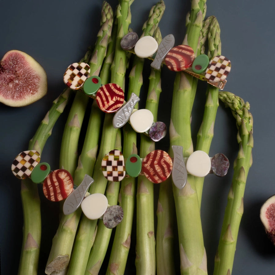 Multiple food themed hair slides sit on asparagus with figs against a dark background