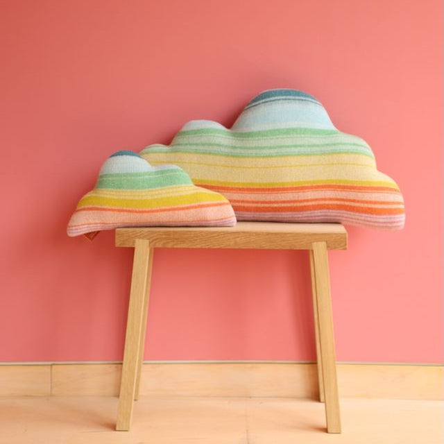 Two cloud-shaped striped knit cushions in pastel rainbow colors resting on a light wood bench against a solid coral-pink wall.