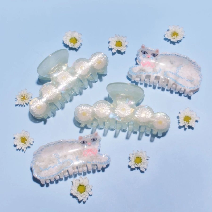 Three translucent iridescent hair claw clips decorated with daisy flowers and a sleeping cat motif, arranged on a light blue background with small daisy props.
