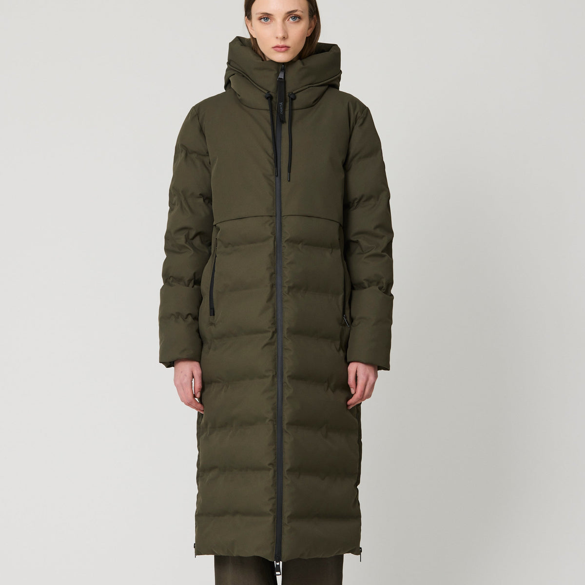 A model stands against a light gray studio background wearing a long, dark olive green puffer parka with a high hood, paired with dark pants and blue sneakers.