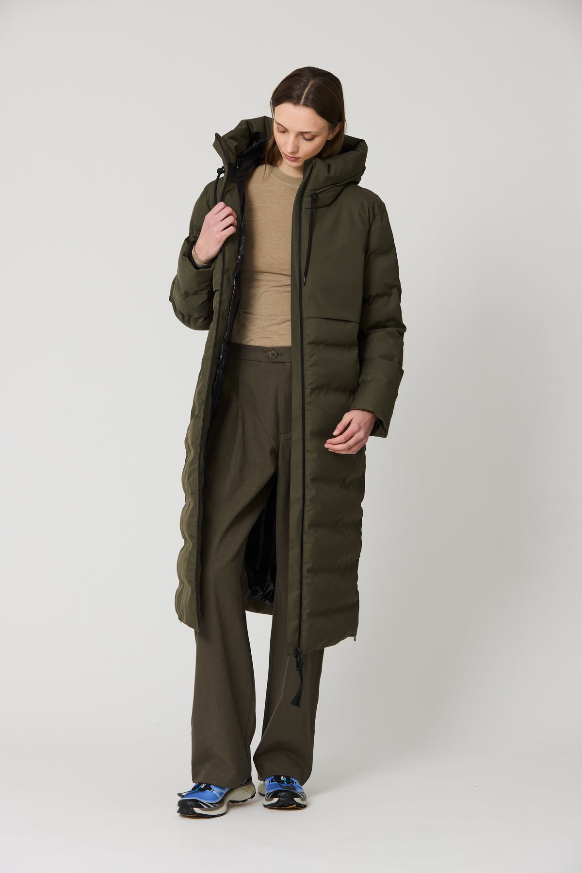 A model wearing a long, dark olive green hooded puffer coat over a beige top and matching dark olive pants, standing against a light gray studio background.