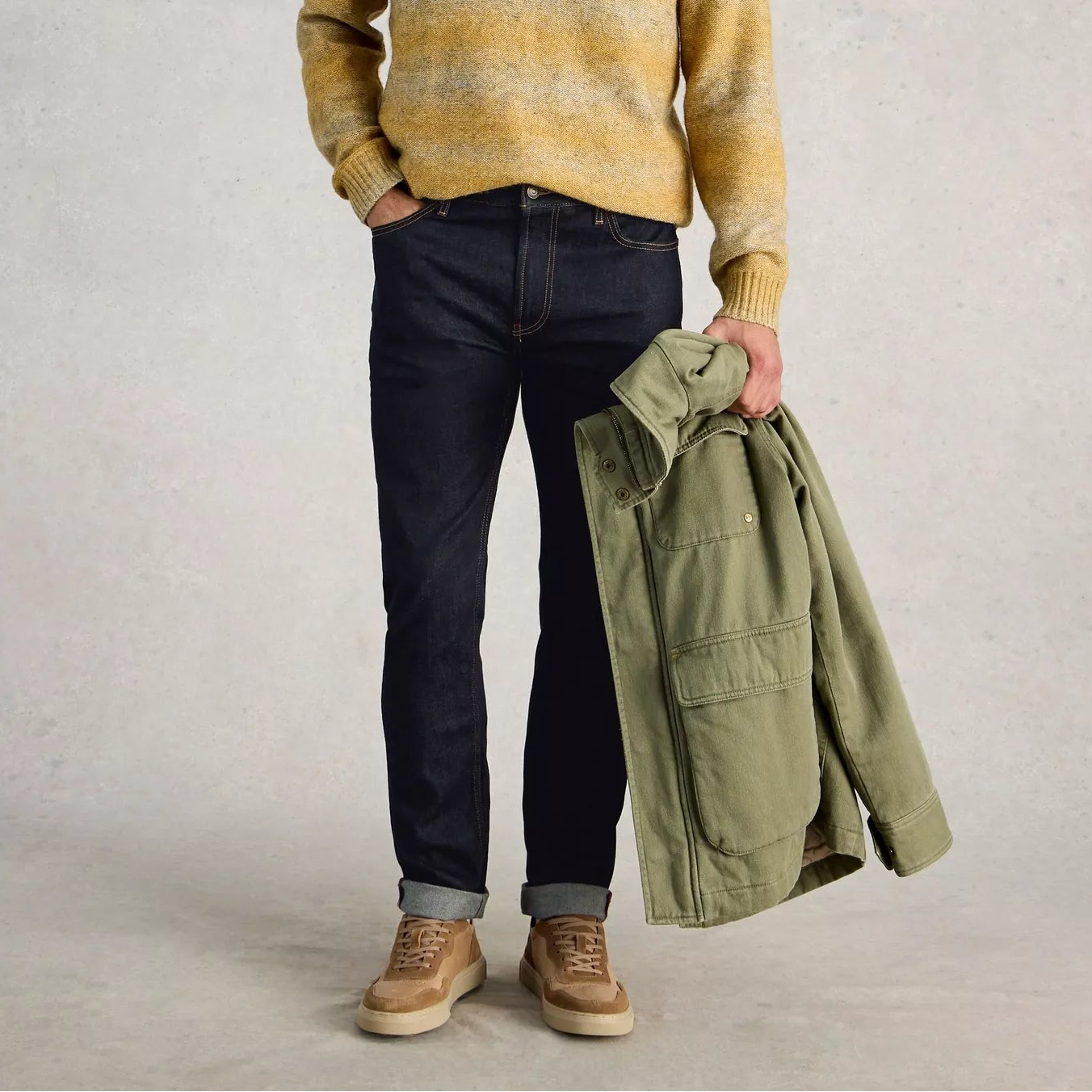 Man wearing a yellow striped sweater, dark jeans, and holding a green jacket against a plain background