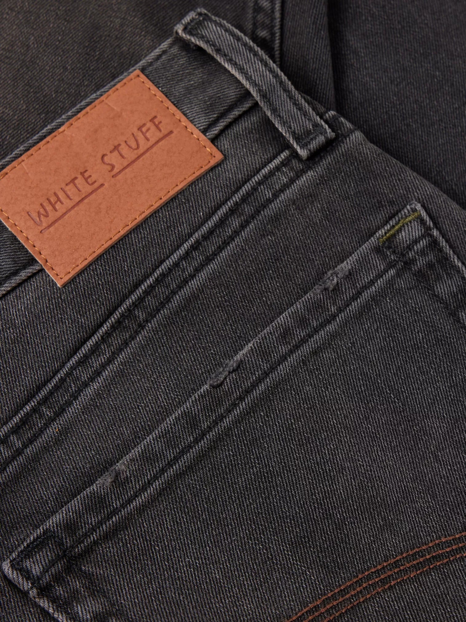 Close-up of dark gray denim jeans back pocket with a brown leather "White Stuff" label.