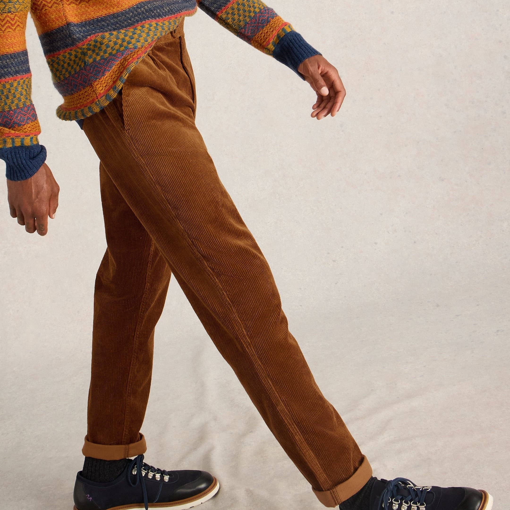 Person wearing a patterned sweater, brown pants, and navy shoes on a neutral background