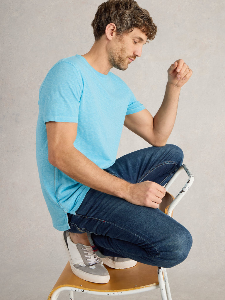 Man in light blue short-sleeved shirt and dark wash jeans sits on a small chair, wearing gray and white sneakers.