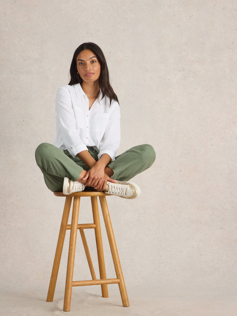 Woman sitting on a wooden stool wearing a white shirt and green pants against a beige background