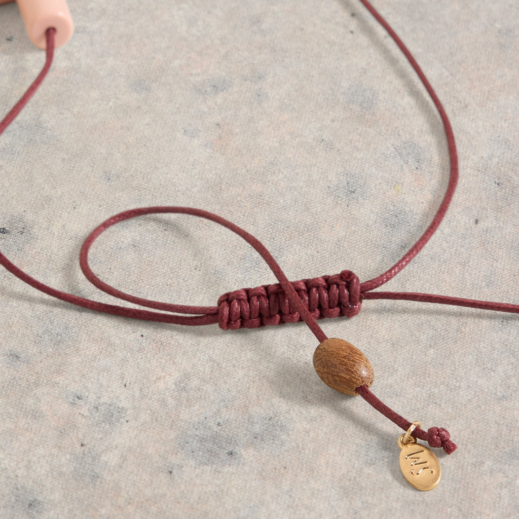 Burgundy cord necklace with a knotted section, dark wood bead, and small gold brand tag.