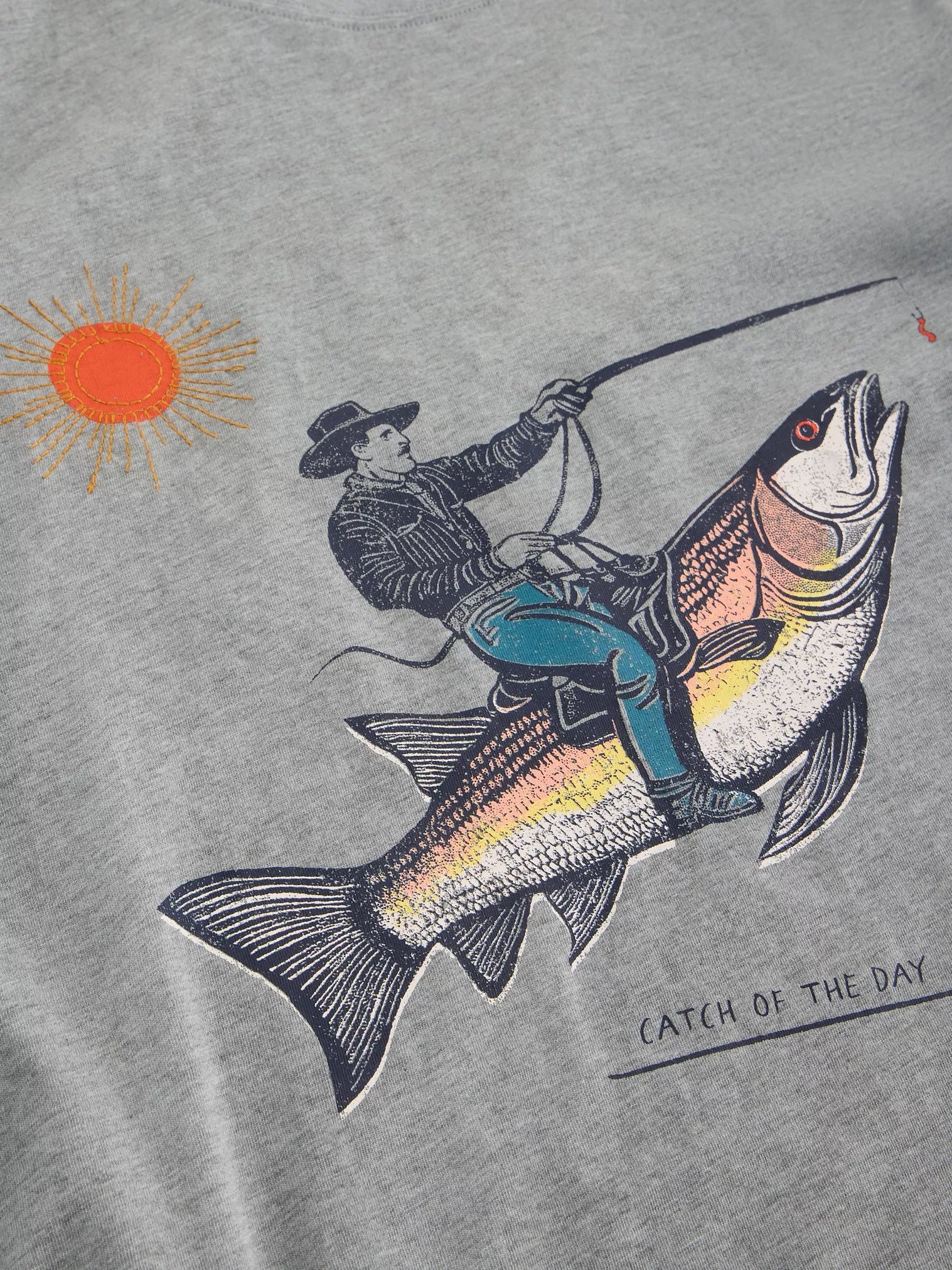 Gray t-shirt featuring a graphic print of a cowboy riding a large fish, with a sun in the corner and "Catch of the Day" text
