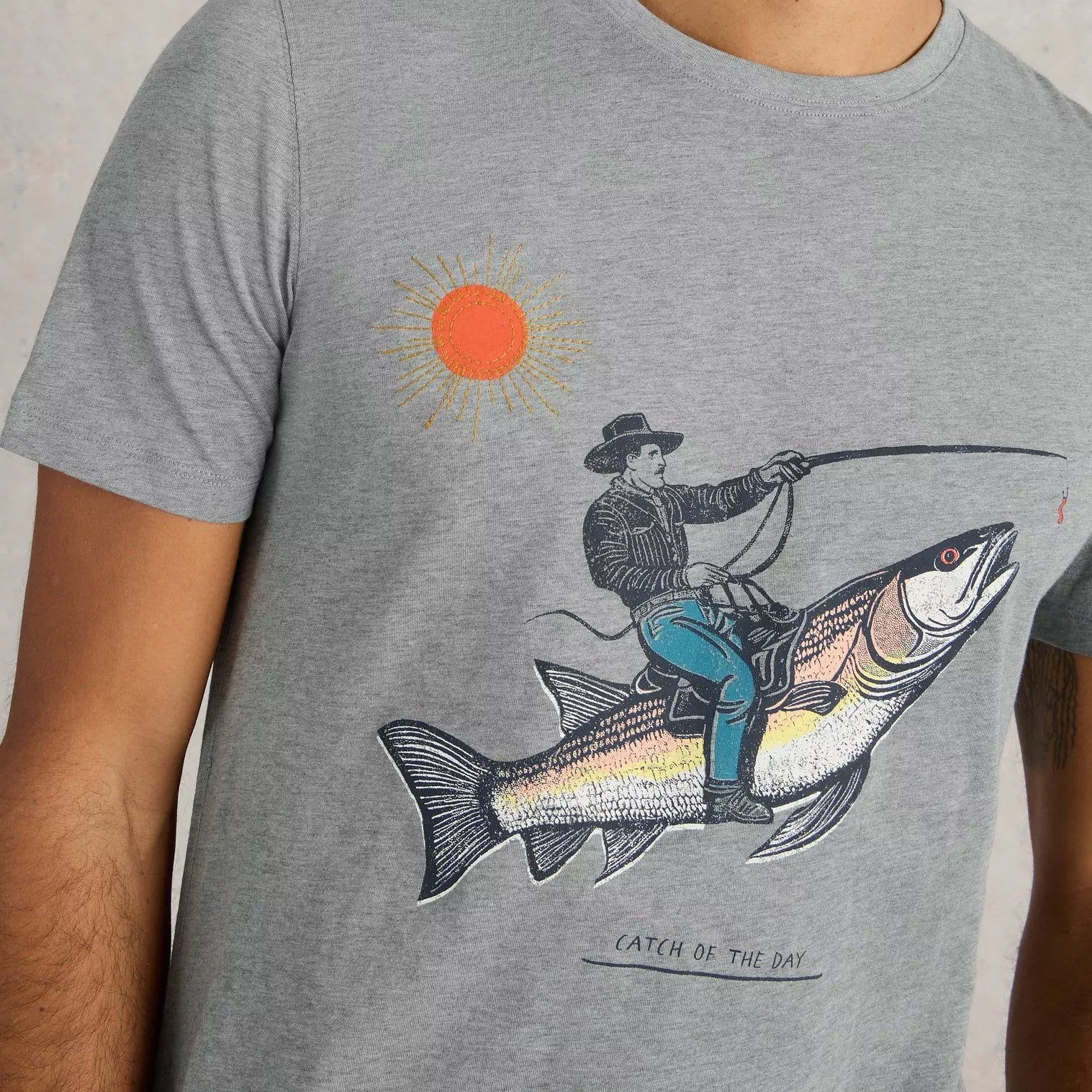 Gray t-shirt featuring a graphic print of a cowboy riding a large fish under a stylized sun.