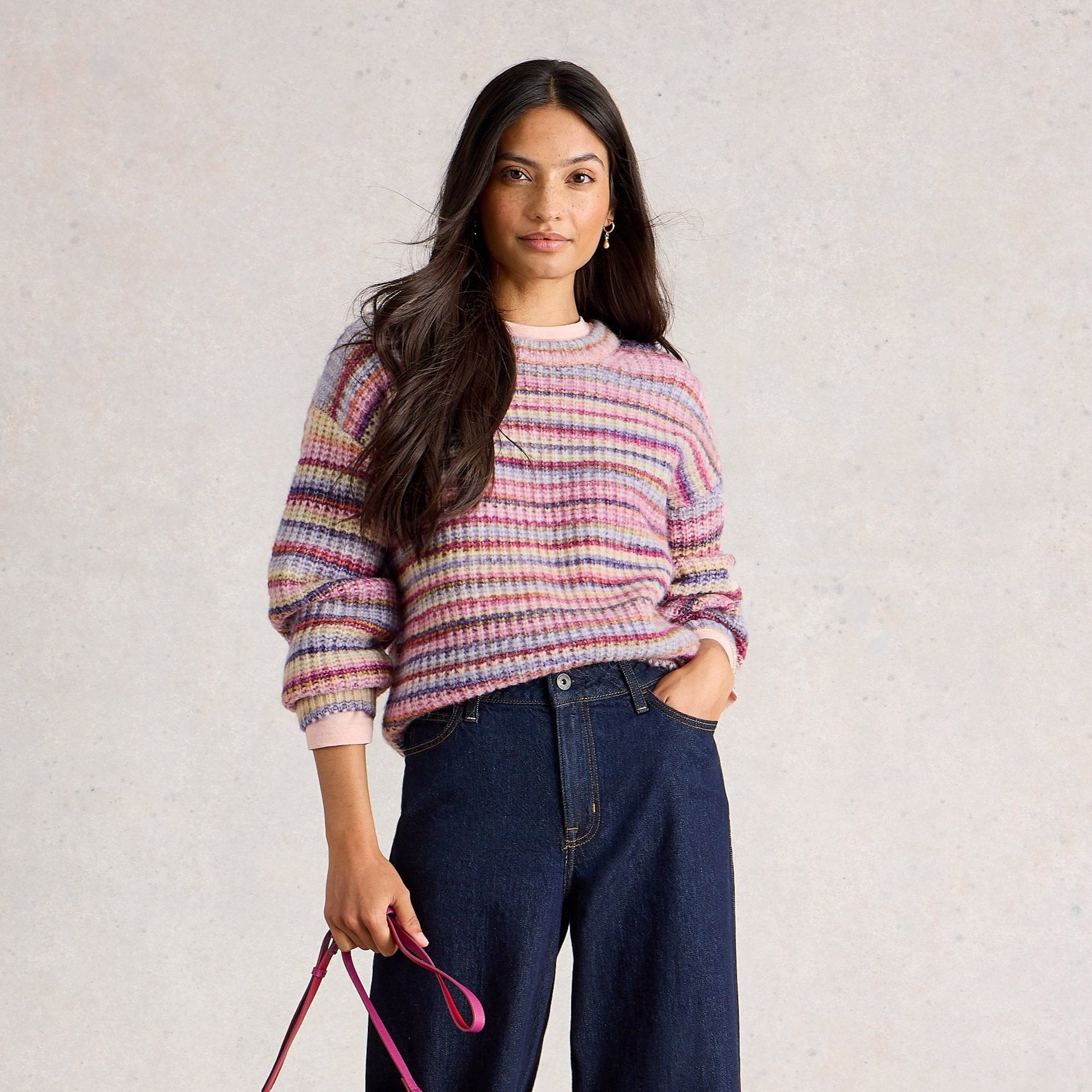 Woman wearing a striped sweater and blue jeans, holding a red handbag on a plain background