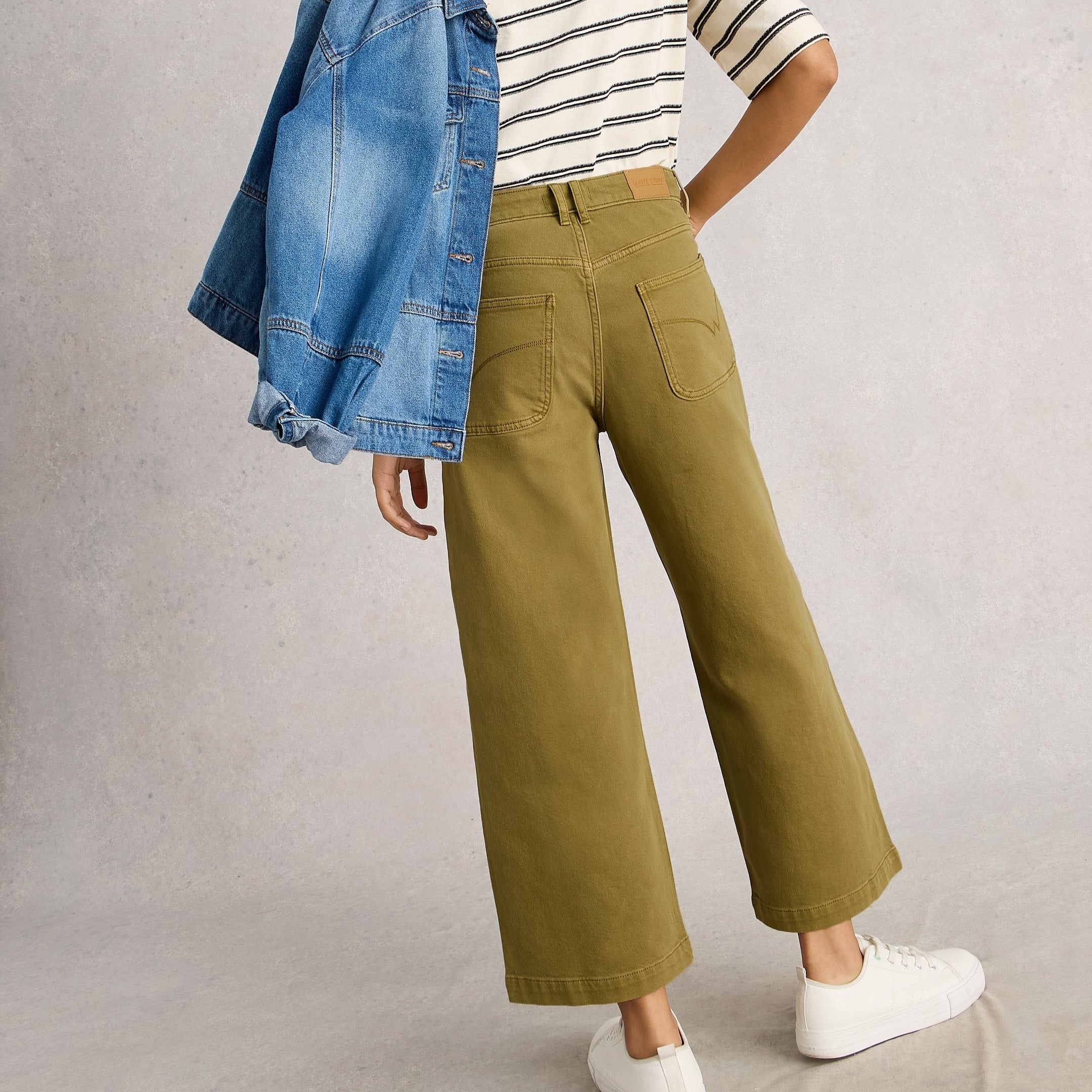 Person wearing a striped shirt, green pants, and white shoes with a denim jacket over one shoulder on a plain background.