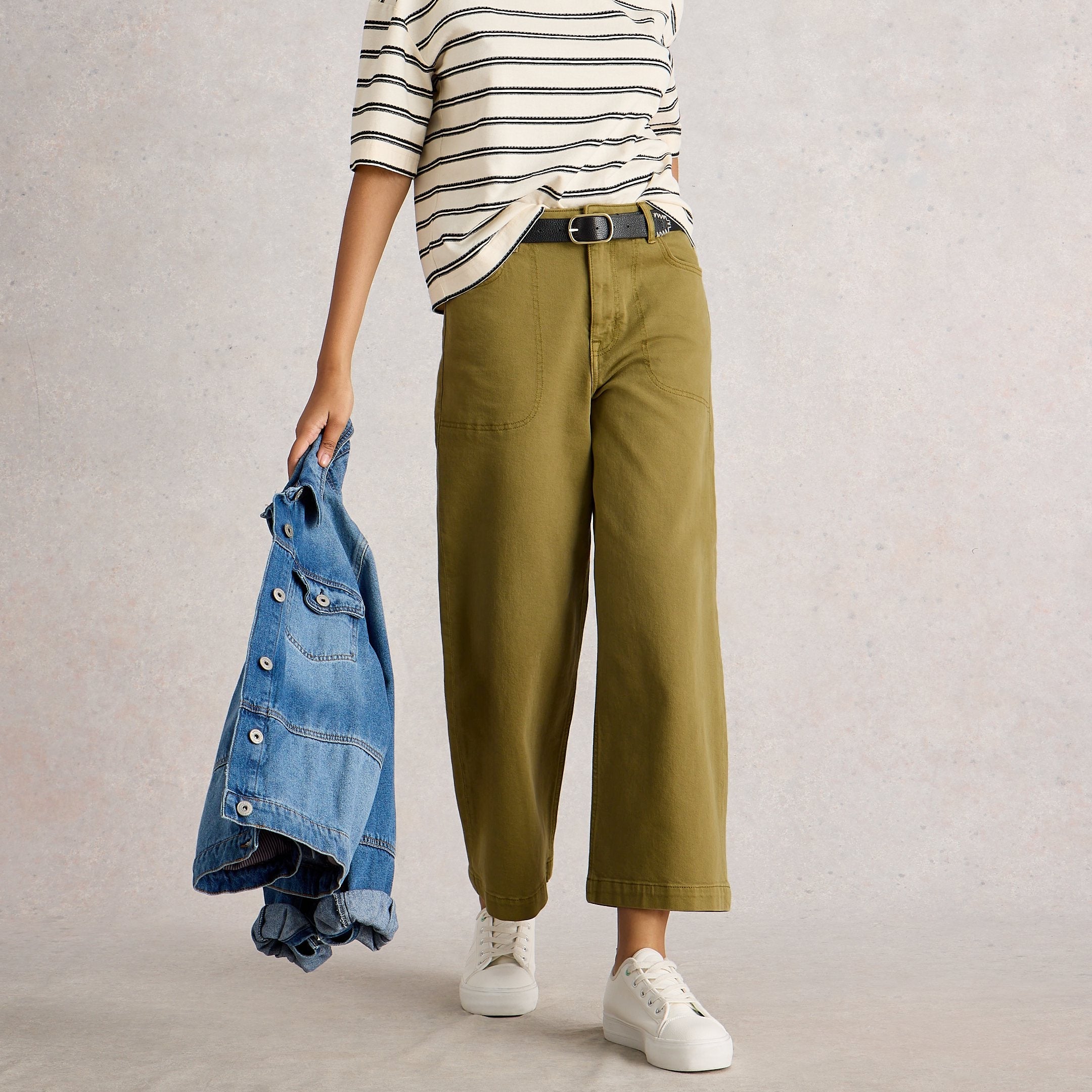 Woman holding a denim jacket, wearing a striped shirt and green pants, on a plain background