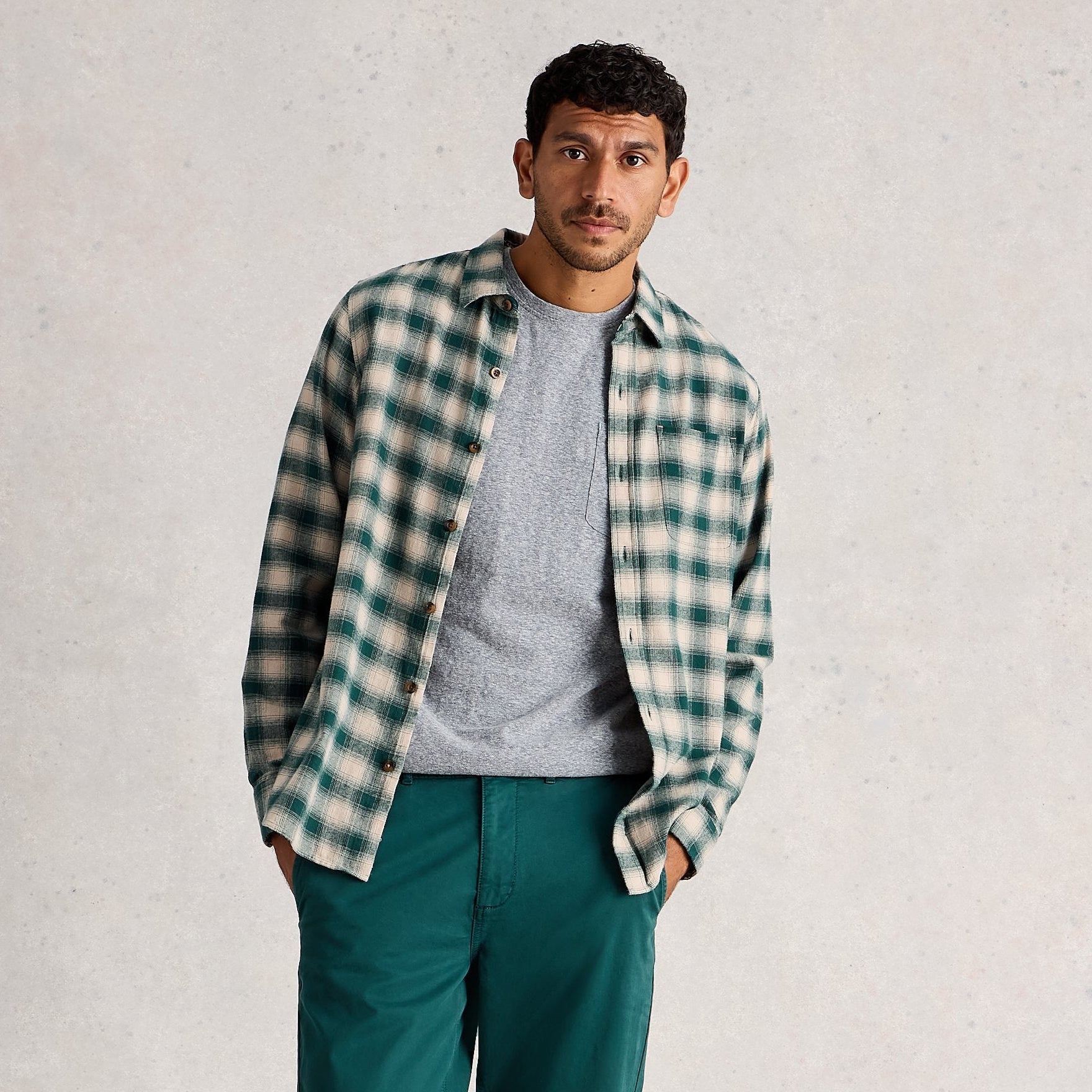 Man wearing a plaid shirt, gray t-shirt, green pants, and white sneakers on a plain background