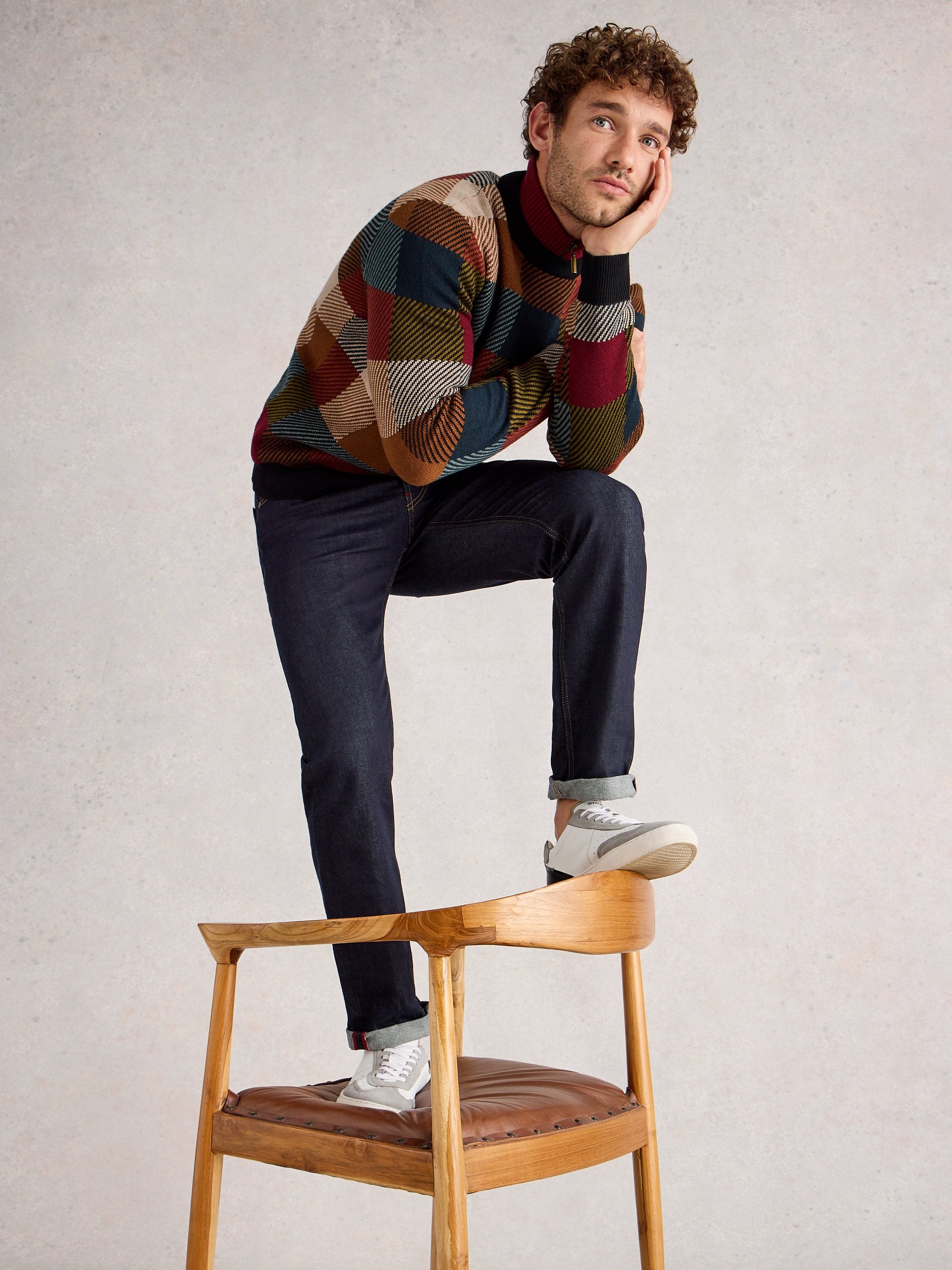 Man sitting on a wooden chair wearing a patterned sweater and dark jeans.